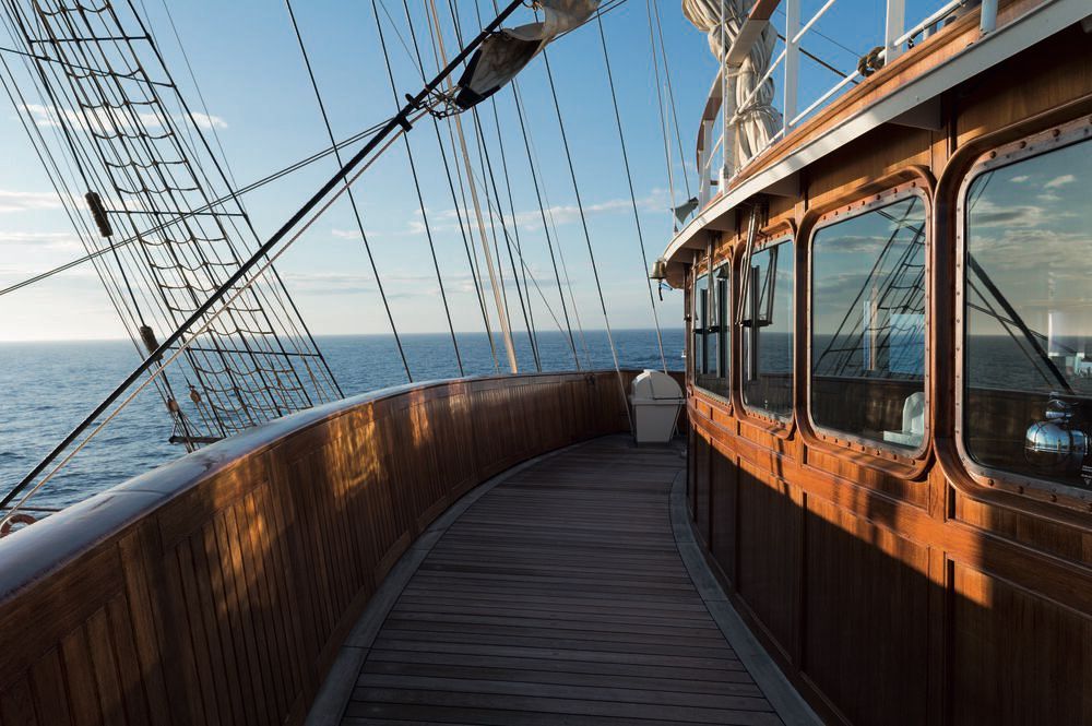 Teakholzdeck entlang des Deckshauses mit Fenstern der Kommandobrücke, Holzreling und Takelage eines Großseglers auf See.