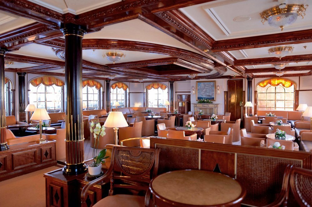 Klassische Lounge mit aufwendiger Mahagoni-Holzvertäfelung, Ledersessel und Bogenfen­stern auf einem Luxuskreuzfahrtschiff
