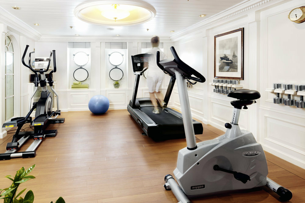 Klassischer Fitnessraum der Sea Cloud II mit weißer Holzvertäfelung, runden Bullaugen, Laufband und historischen Segelschiff-Kunstdrucken