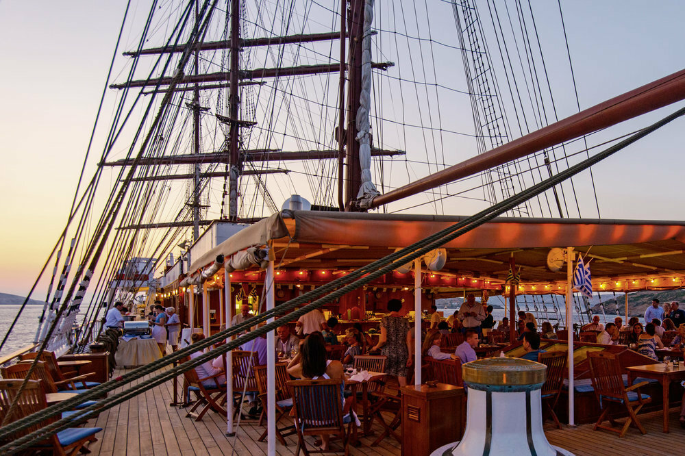 Abendessen auf dem Deck eines Großseglers bei Sonnenuntergang mit Masten, Takelage und griechischer Küste im Hintergrund