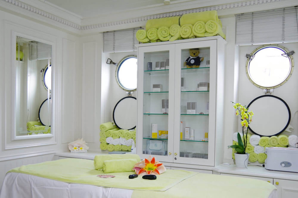 Heller Spa-Behandlungsraum der Sea Cloud II mit Massageliege, gerollten Handtüchern und runden Bullaugenfenstern.