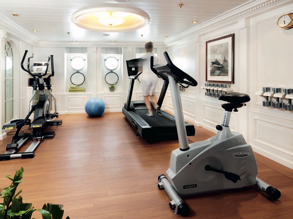 Fitnessraum an Bord mit Laufband, Crosstrainer, Ergometer, Hantelablage und Gymnastikball vor weißer Wandvertäfelung.