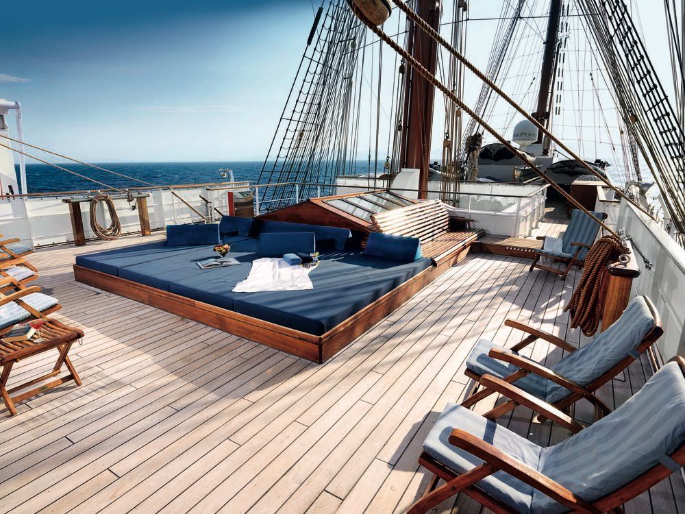 Sonnendeck eines Großseglers mit großer gepolsterter Liegeinsel, Holzliegestühlen, Teakdeck und Takelage mit Blick aufs offene Meer.