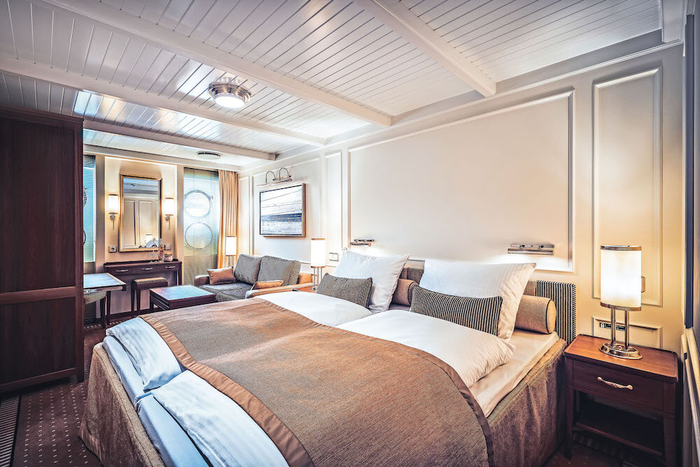 De-Luxe-Außenkabine mit Doppelbett, Bullaugenfenstern, Schminktisch und klassischer Ausstattung.