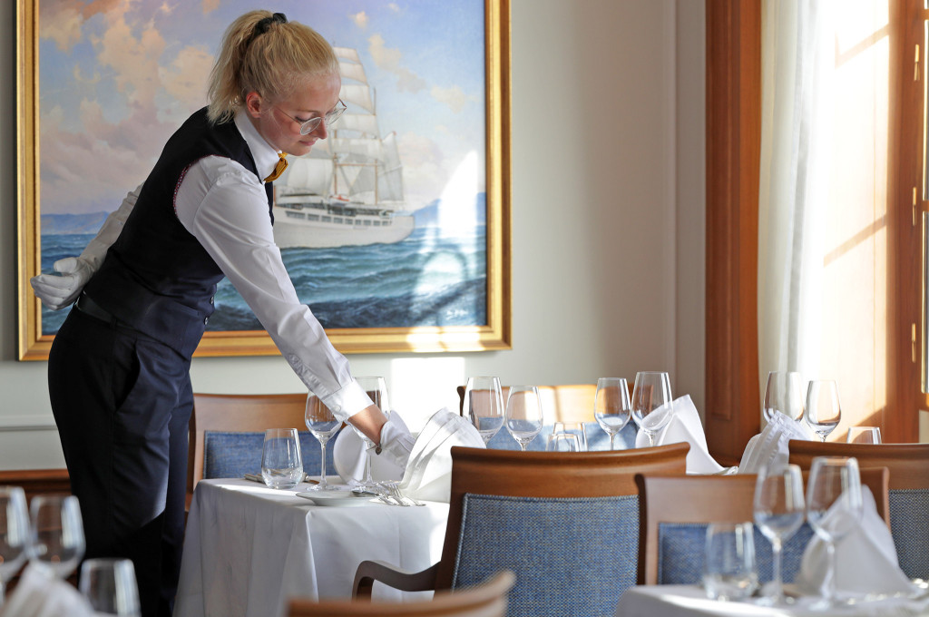 Servicemitarbeiterin deckt einen Tisch im hellen Restaurant der Sea Cloud Spirit vor einem Gemälde des Segelschiffs.