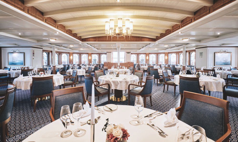 Bordrestaurant eines Großseglers mit runden, weiß eingedeckten Tischen, Holzstühlen, Bogenfenstern und Deckenleuchter.