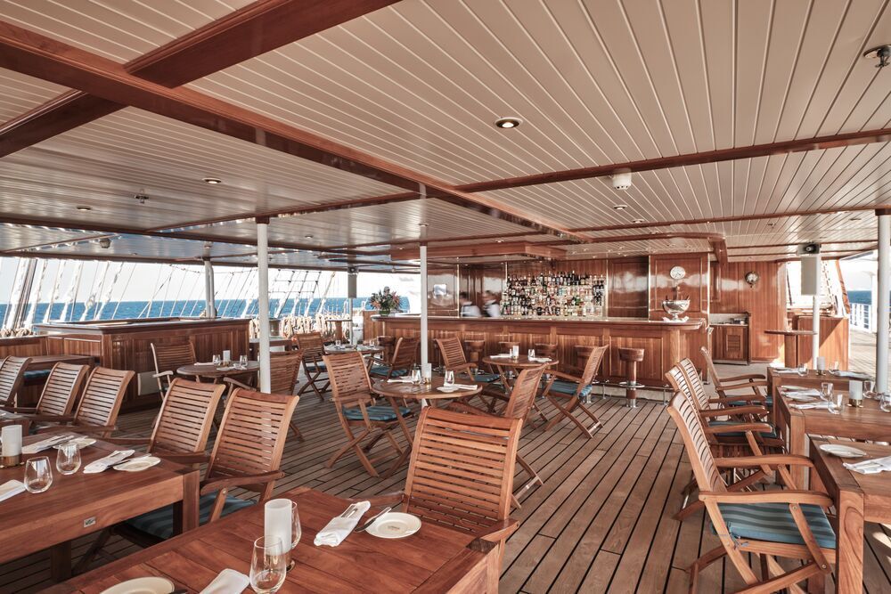 Überdachte Bar an Deck eines Großseglers mit Bartresen, Barhockern, Holztischen und Klappstühlen mit Blick aufs Meer.