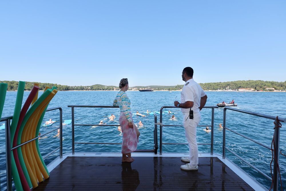 Gast und Crewmitglied auf der Badeplattform am Heck, während Badende in der Bucht vor der Küste schwimmen.