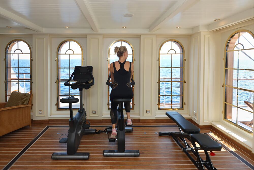 Frau trainiert auf einem Heimtrainer im Fitnessbereich mit Bogenfenstern und Blick auf das Meer.