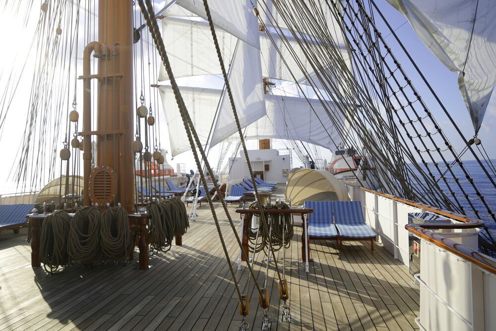 Sonnendeck eines Großseglers mit gestreiften Sonnenliegen auf Teakdeck, aufgeschossenem Tauwerk, Mast und gesetzten Segeln.