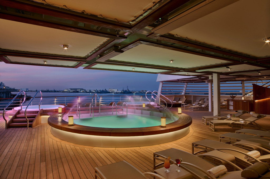 Pooldeck der Seabourn Ovation bei Dämmerung mit rundem Whirlpool, Liegestühlen und Hafenpanorama im Hintergrund.