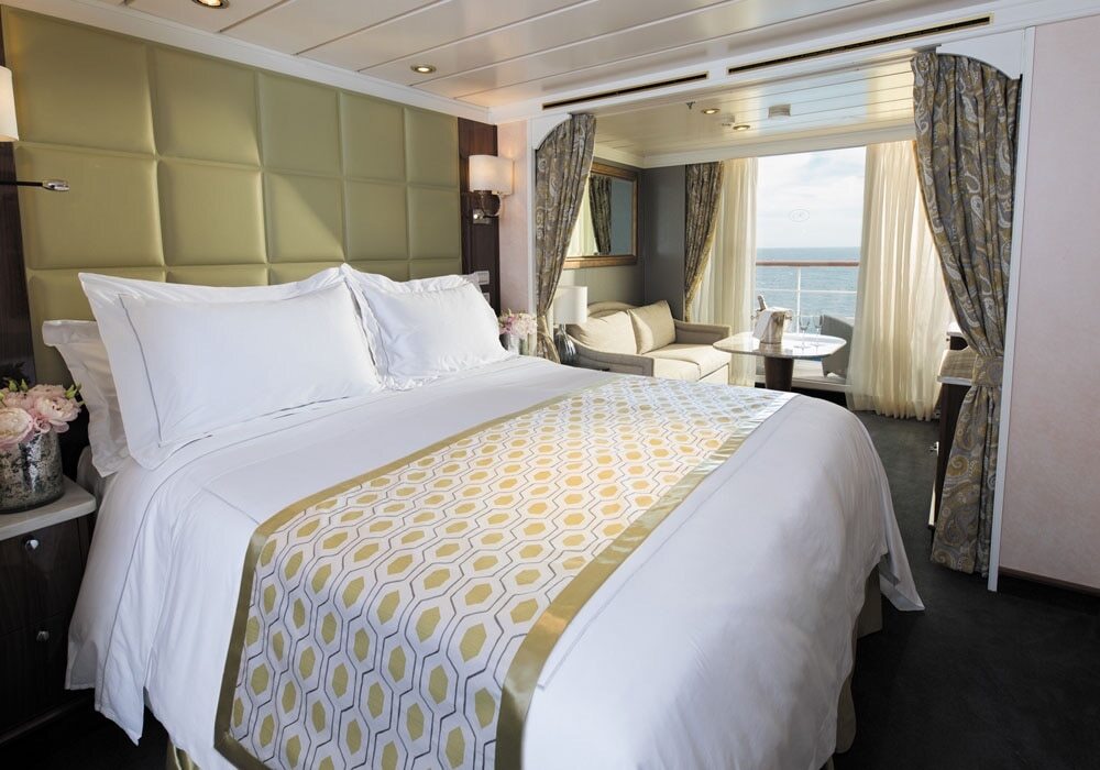 Penthouse Suite mit gepolstertem Kopfteil, weißen Bettwaren mit goldenem Läufer, Sitzecke und Balkon mit Meerblick auf der Seven Seas Mariner