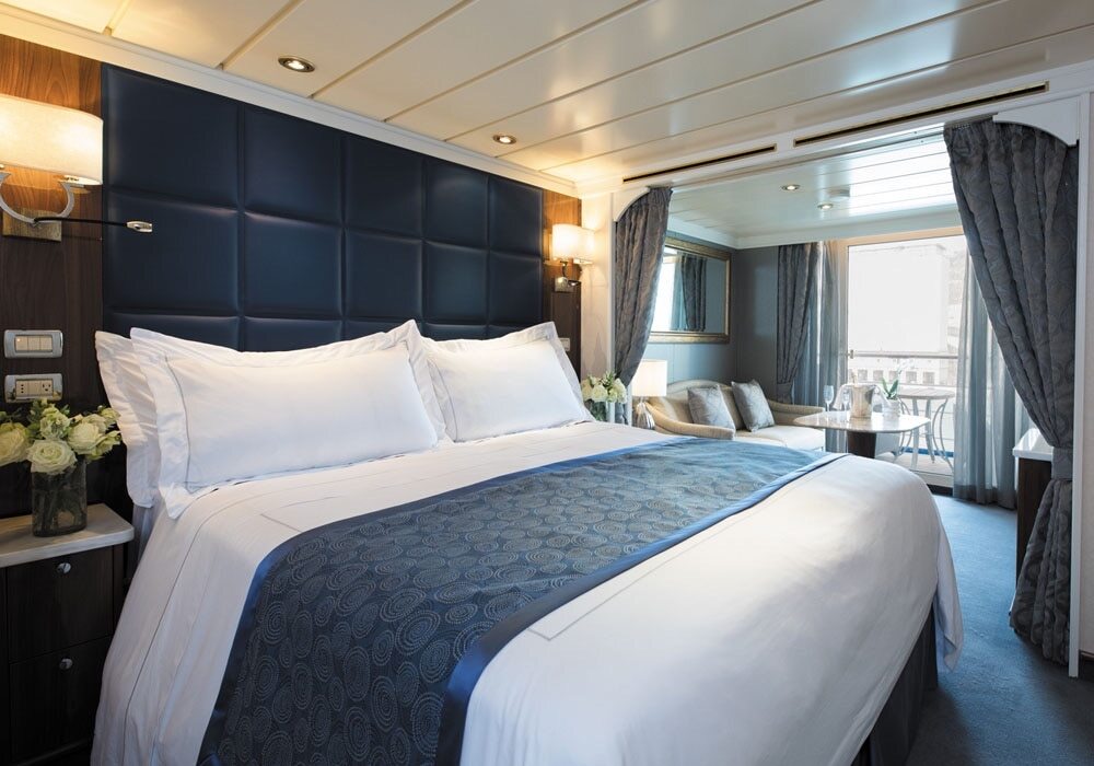 Deluxe Veranda Suite mit marineblauem Kopfteil, weißen Bettwaren, Sitzecke und Balkon mit Meerblick auf der Seven Seas Mariner