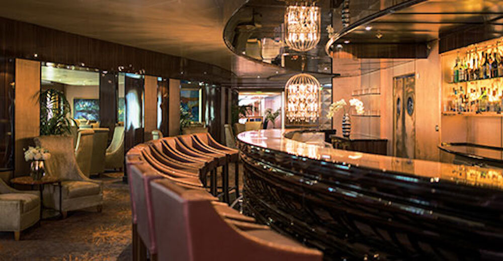 Elegante Bar mit geschwungener schwarzlackierter Theke, Kristallleuchter und warmem Holzdekor auf einem Luxuskreuzfahrtschiff