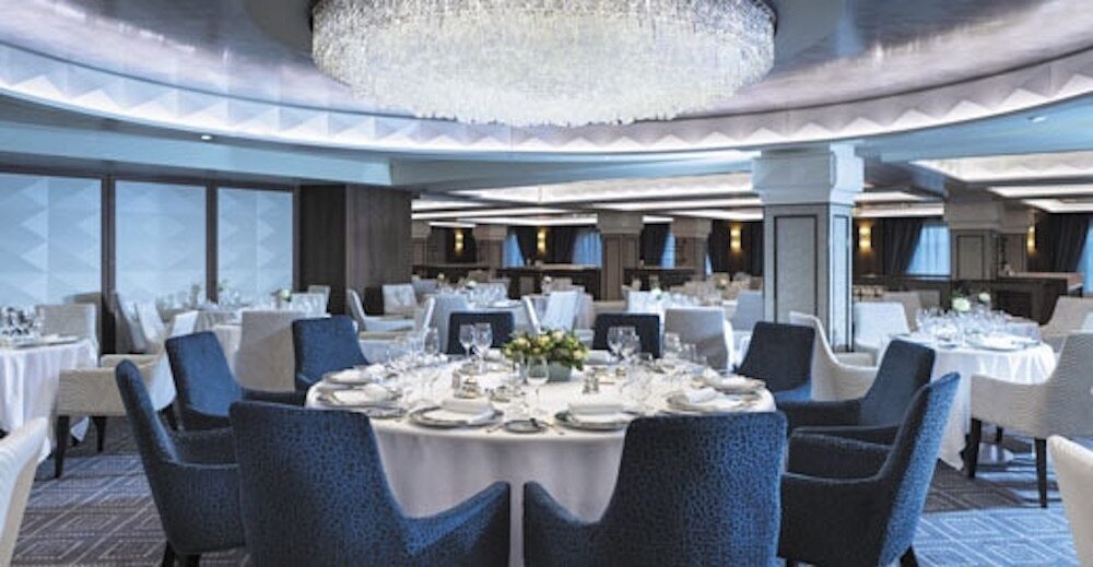 Elegantes Compass Rose Hauptrestaurant mit großem Kristallkronleuchter, runden Tischen und blauen Sesseln auf der Seven Seas Mariner