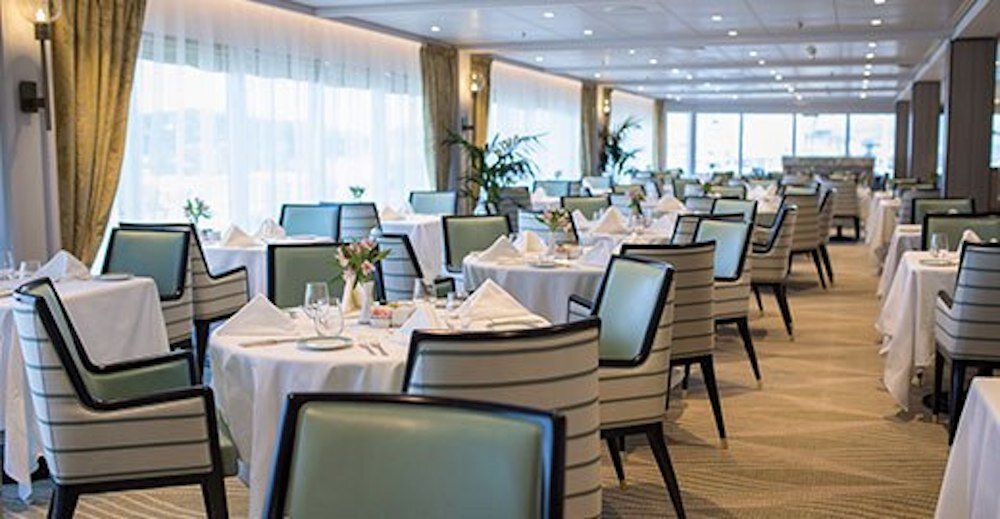 Helles Restaurant mit runden Tischen, weißen Tischdecken und großen Panoramafenstern mit Meerblick auf einem Luxuskreuzfahrtschiff