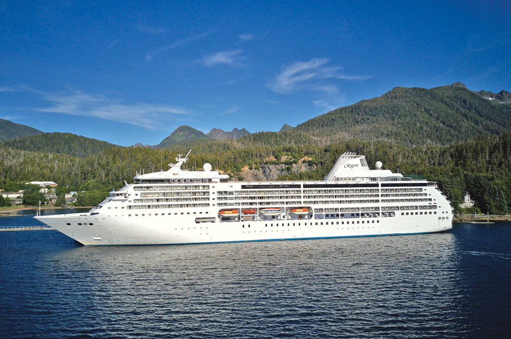 Die Seven Seas Mariner liegt vor bewaldeten Bergen in einem ruhigen Fjord, fotografiert von der Seite bei strahlendem Sonnenschein.