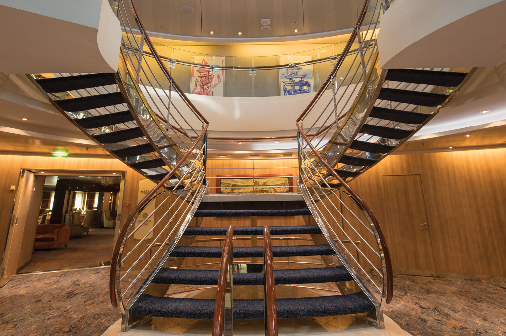 Atrium der Seven Seas Mariner mit geschwungener Doppeltreppe, Holzvertäfelung und großzügiger Deckenhöhe über mehrere Etagen.
