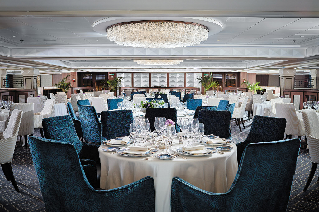Hauptrestaurant Compass Rose mit imposantem Kristallkronleuchter und festlich gedeckten Tischen mit blauen Polsterstühlen an Bord der Seven Seas Navigator.