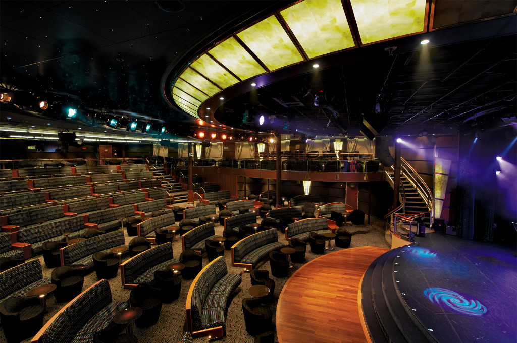 Theater an Bord der Seven Seas Navigator mit gepolsterten Sitzreihen, beleuchteter Bühne und kunstvoller Deckengestaltung.