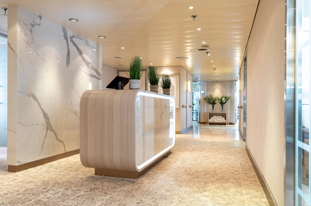Heller Spa-Empfang mit geschwungenem Holztresen, eleganter Marmorwand und Grünpflanzen an Bord der Seven Seas Navigator.