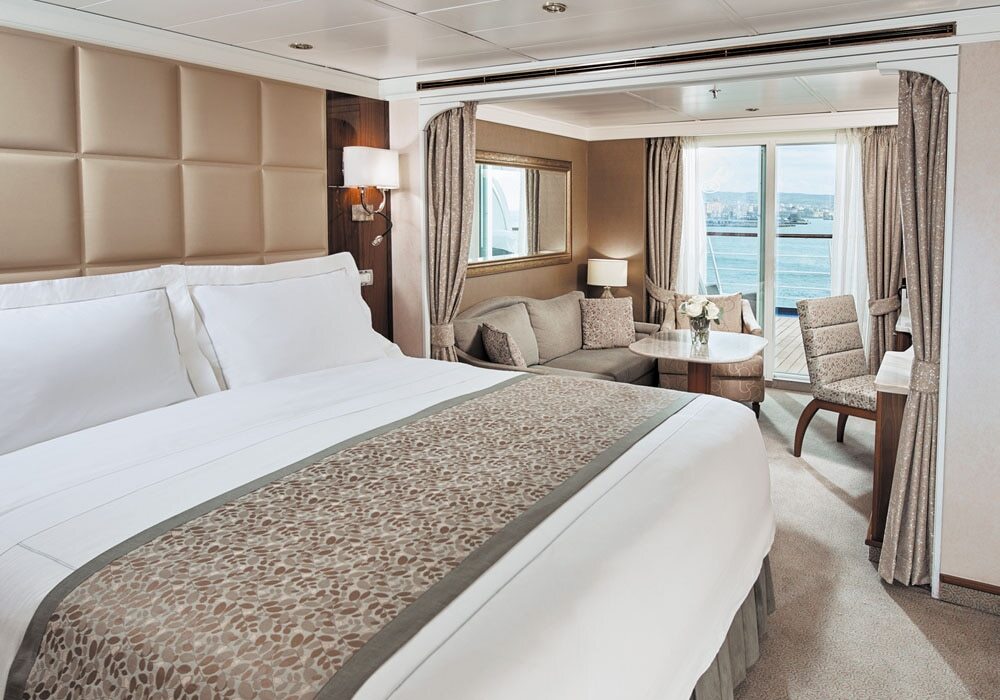Penthouse Suite mit Doppelbett, Polsterkopfteil, Sitzecke und Balkon mit Hafenstadtblick auf einem Hochseekreuzfahrtschiff