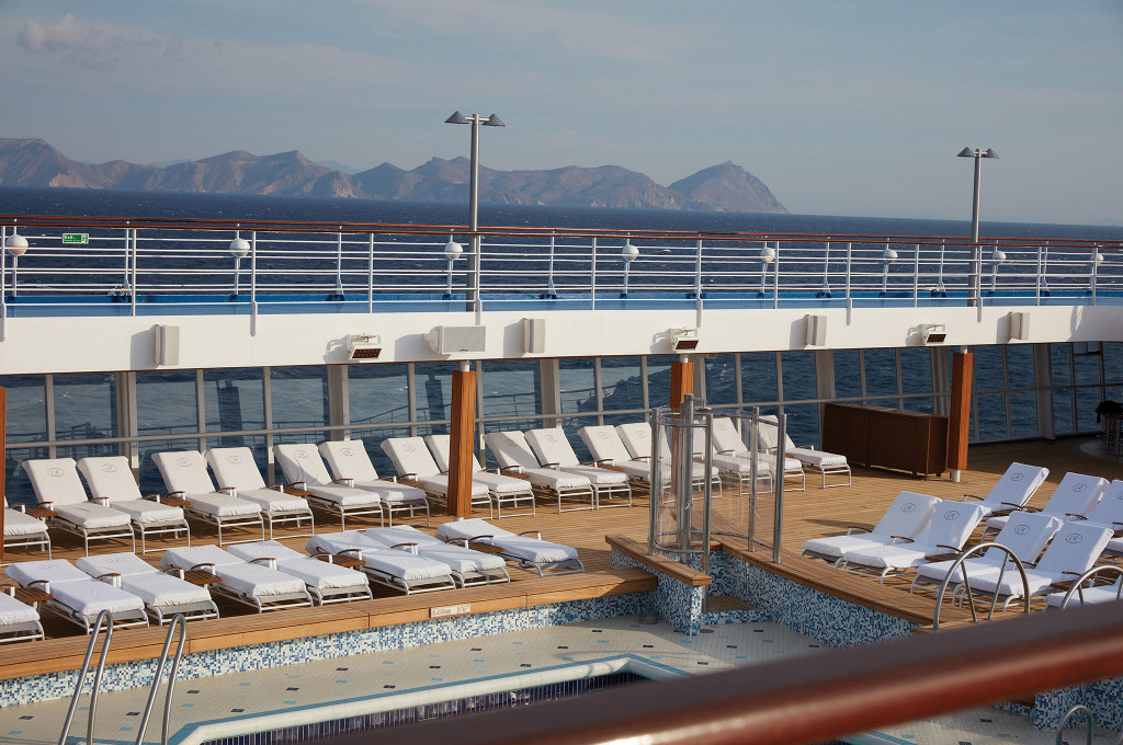 Pooldeck mit Außenpool, weißen Liegestühlen und Blick auf eine Bergküste am Horizont.