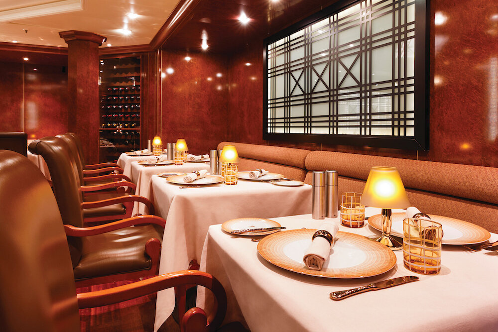 Intimes Spezialitätenrestaurant mit weißen Tischdecken, Lederstühlen, Kerzenleuchtern und Weinregal auf einem Luxuskreuzfahrtschiff