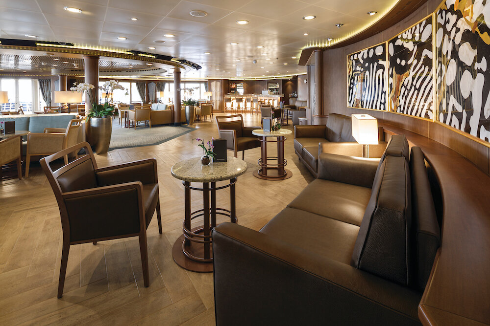 Elegante Panorama-Lounge mit Ledersofas, Marmortischen, Parkettboden und Gemälden auf einem Luxuskreuzfahrtschiff