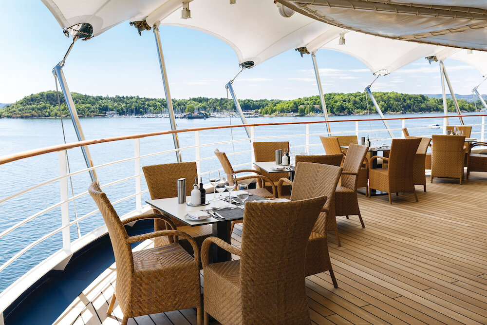 Überdachte Außenterrasse eines Kreuzfahrtschiffes mit Rattanstühlen und Blick auf grüne Küstenlandschaft und Fjord