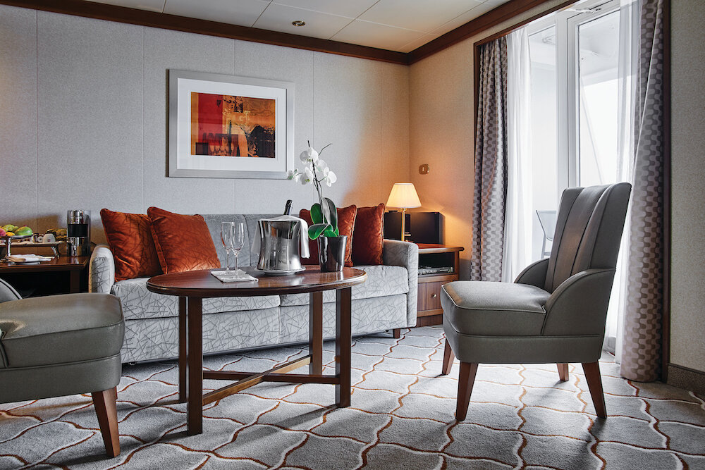 Wohnbereich einer Suite mit gepolstertem Sofa, Ledersesseln, ovalem Couchtisch mit Champagnerkühler und Orchidee auf einem Kreuzfahrtschiff