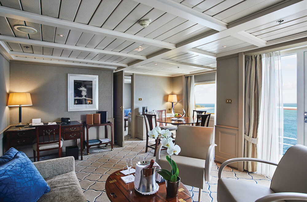 Klassischer Suite-Wohnbereich mit Sofa, Sesseln, Esstisch, Orchidee, Champagner und Panoramafenster mit Meerblick