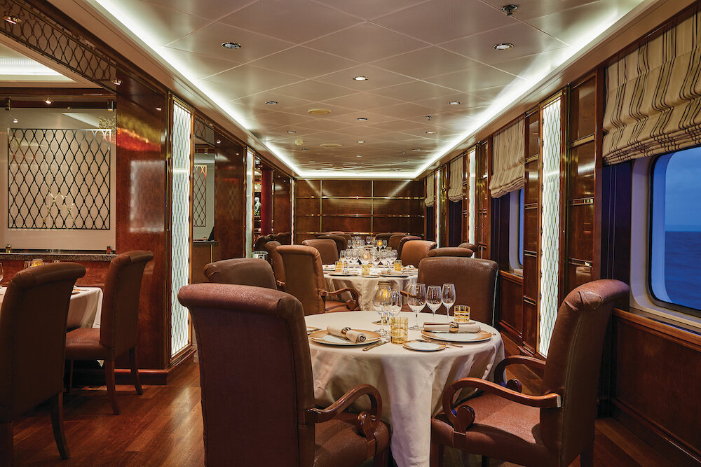 Elegantes Spezialitätenrestaurant mit dunkler Holzvertäfelung, gedeckten Rundtischen und Lederpolstersesseln auf einem Kreuzfahrtschiff