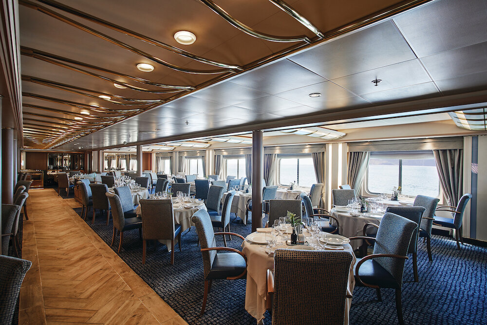 Klassisches Hauptrestaurant mit weißen Tischdecken, gemusterten Stühlen und Panoramafenstern auf einem Expeditionsschiff