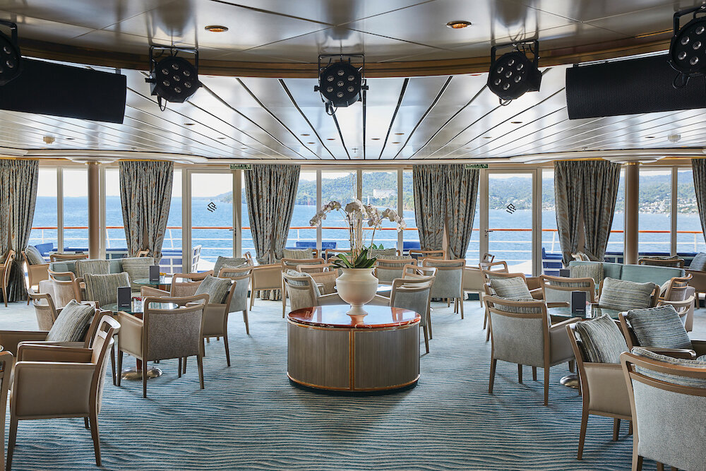Elegante Panoramalounge mit Sesseln, Rundetischen und großen Fenstern mit Meerblick auf einem Expeditionskreuzfahrtschiff