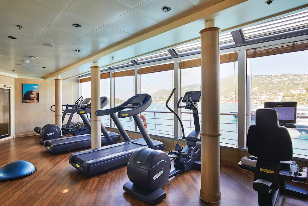 Fitnessraum der Silver Wind mit Laufbändern, Ellipsentrainer, Holzsäulen und Panoramafenstern mit Blick auf Hafen und Küste