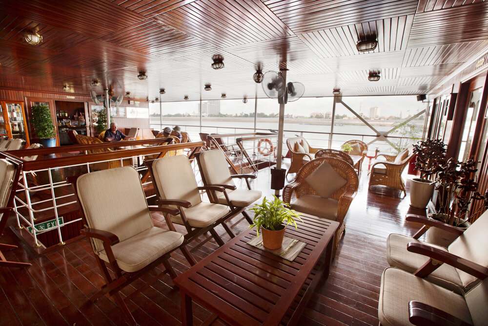 Überdachte Lounge mit Holzdeck, Korbsesseln und Liegestühlen sowie Bartresen im Hintergrund und Blick auf den Fluss.