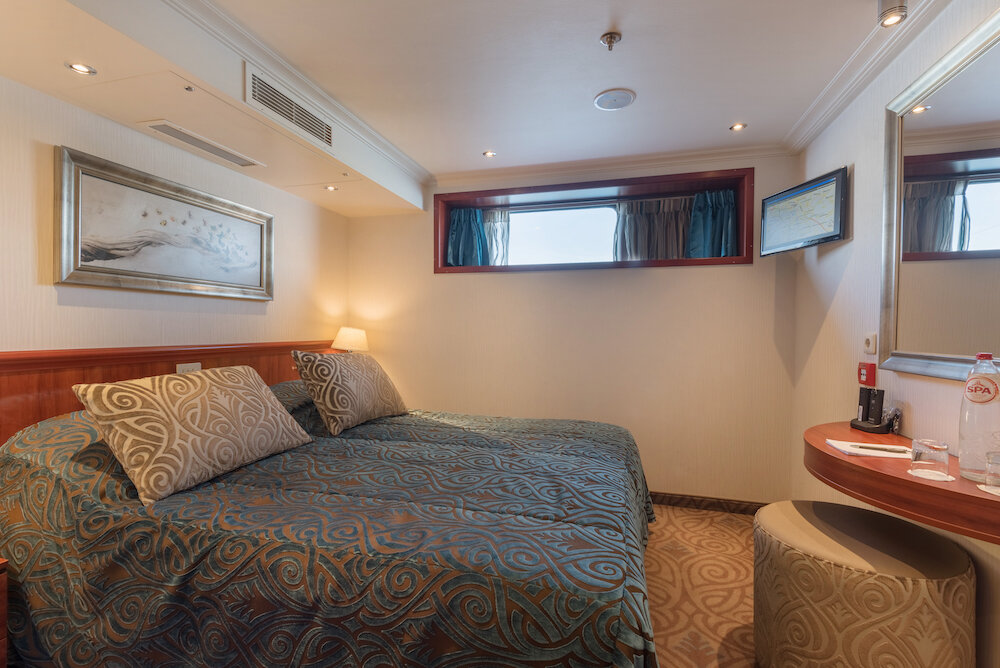 Zweibettkabine auf dem Hauptdeck mit zusammengestelltem Doppelbett, gemusterter Tagesdecke, Schminktisch und Fenster