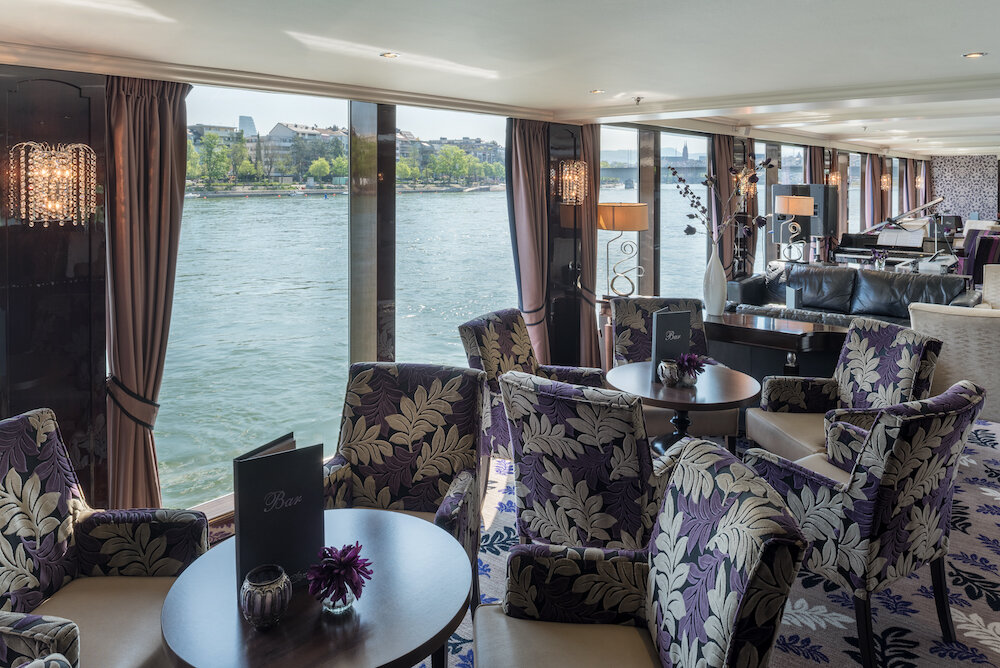 Panorama-Salon eines Flusskreuzfahrtschiffes mit gemusterten Sesseln, Bistrotischen, Klavier und Fensterfront zum Fluss.