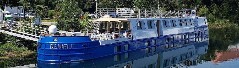 Die Péniche Daniele liegt an einem von Bäumen umgebenen Kanalufer, auf dem Vorschiff ein beschatteter Sitzbereich.