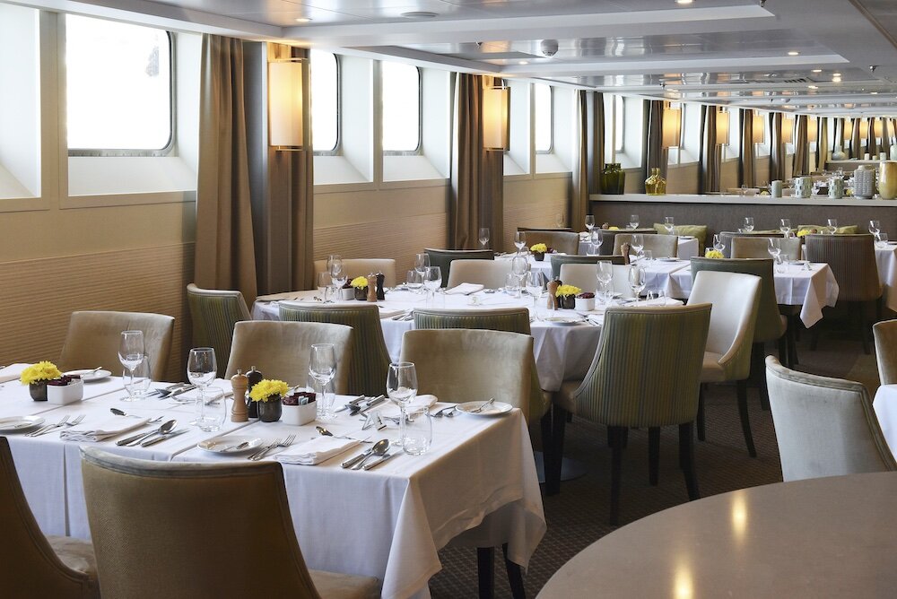 Restaurant an Bord mit eingedeckten Tischen, Polsterstühlen und einer Fensterreihe.