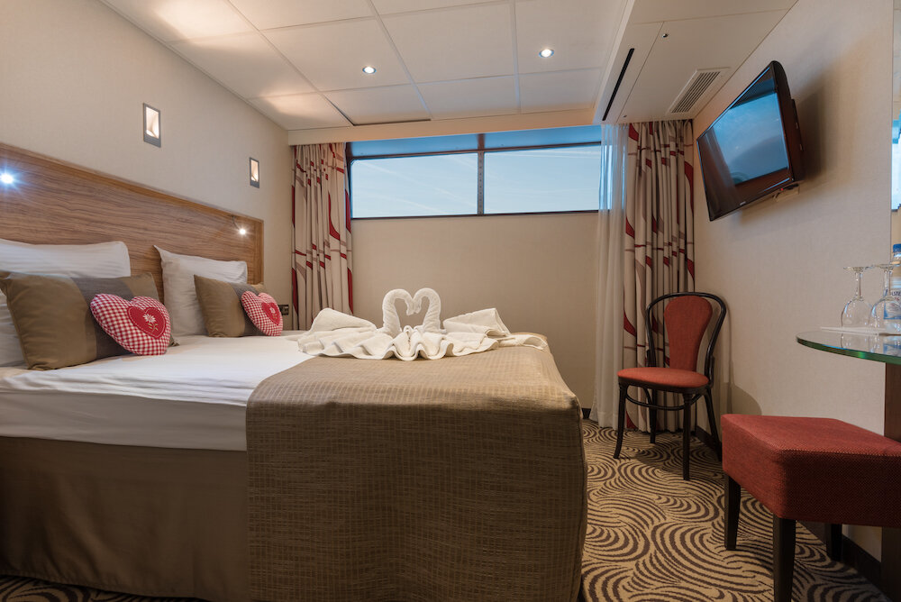 Außenkabine auf dem Hauptdeck eines Flusskreuzfahrtschiffs mit Doppelbett, Fenster, Wandfernseher und Sitzecke.