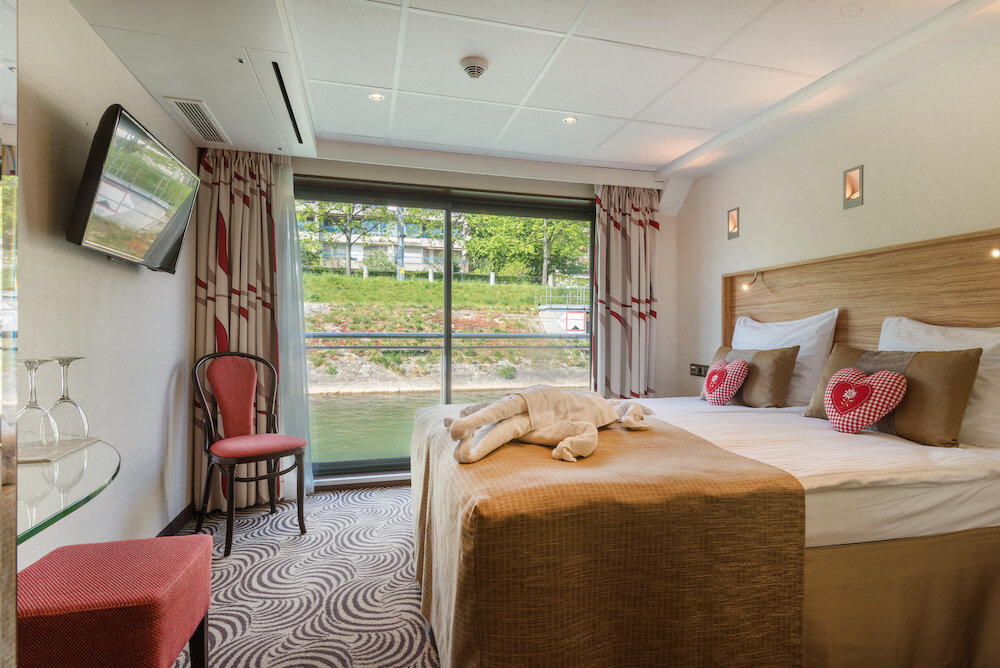 Kabine mit französischem Balkon auf einem Flusskreuzfahrtschiff, Doppelbett und Blick durch die Schiebetür auf das Ufer.