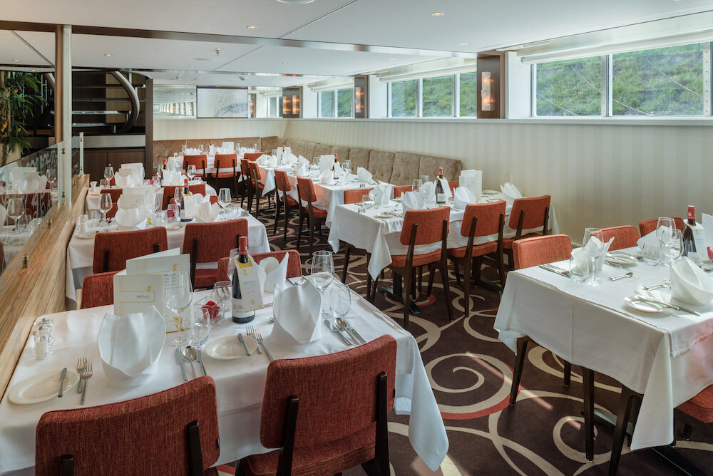 Restaurant auf einem Flusskreuzfahrtschiff mit eingedeckten Tischen, weißen Tischdecken und Fensterreihe zum Ufer.