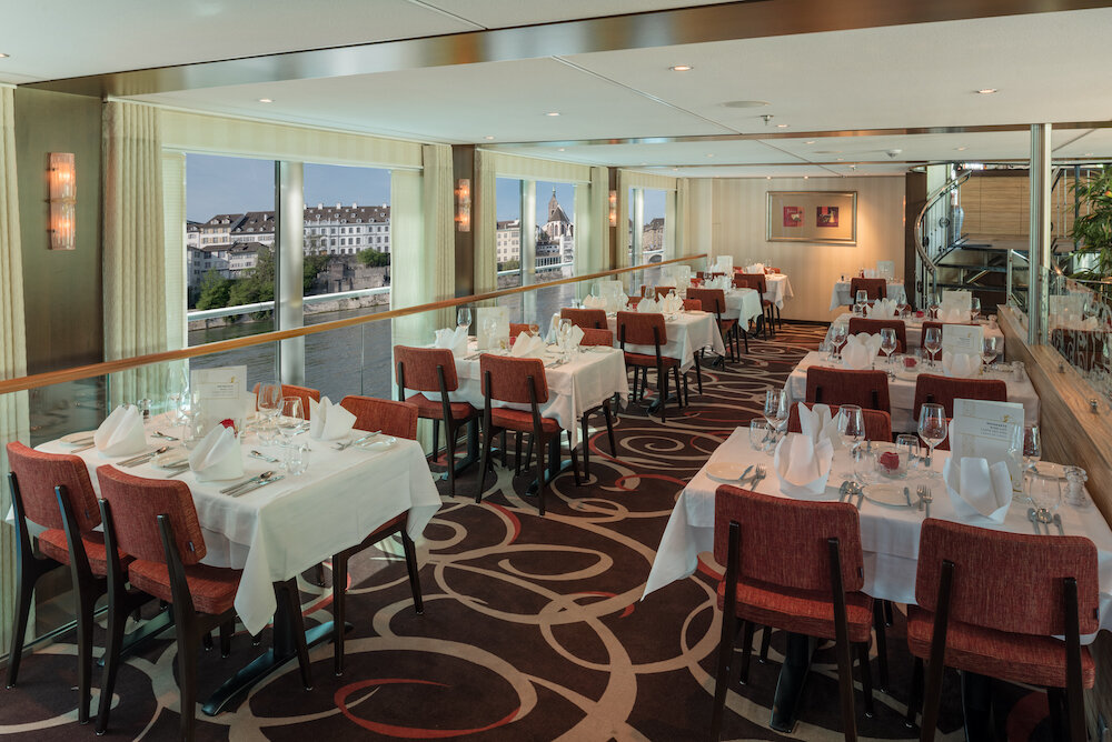 Restaurant eines Flusskreuzfahrtschiffs mit eingedeckten Tischen an Panoramafenstern und Blick auf eine Altstadt am Fluss.