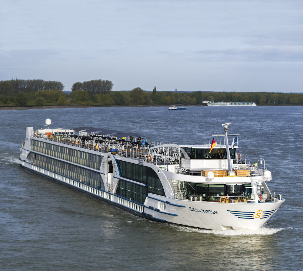 Flusskreuzfahrtschiff in Fahrt auf dem Rhein, Blick auf Bug und Sonnendeck vor bewaldetem Ufer.