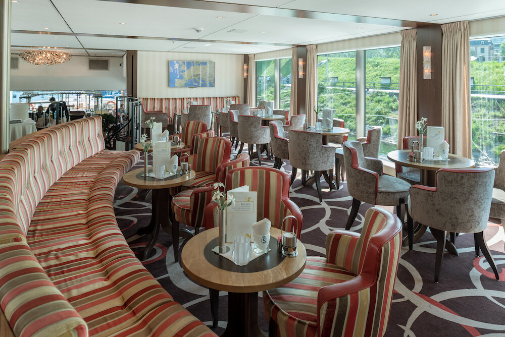 Panorama-Salon eines Flusskreuzfahrtschiffs mit gestreifter Sitzbank, Sesselgruppen an runden Tischen und großen Fenstern.