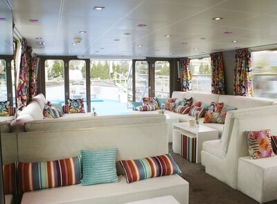 Helle Lounge eines Flusskreuzfahrtschiffes mit weißen Sofas, bunt gestreiften Kissen und Panoramafenstern