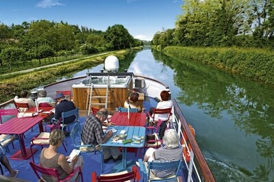 Sonnendeck eines Flusskreuzfahrtschiffes mit Tischen und Stühlen, auf dem Gäste während der Fahrt durch einen von Bäumen gesäumten Kanal sitzen