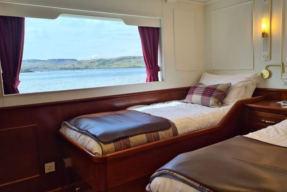 Außenkabine mit Einzelbett, Mahagoni-Holzvertäfelung und breitem Panoramafenster mit Blick auf die schottische Küstenlandschaft.